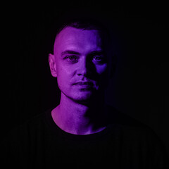 A man stands under vibrant purple lighting, expressing a thoughtful demeanor. The dark background enhances his features, creating an engaging visual contrast