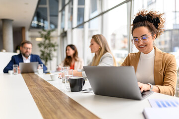 Smiling lawyer reviewing report on her laptop with legal colleagues collaborating in the background during professional meeting