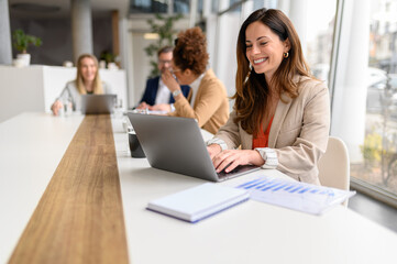 Happy female professional analyzing financial report on laptop while legal colleagues discussing in the background