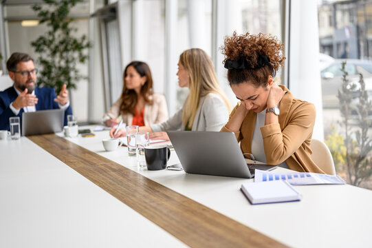 Stressed businesswoman holding her neck in pain and working on laptop while colleagues discussing in meeting