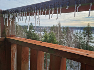 View of a snowy panorama on a sunny day from a wooden balcony. Melting icicles hanging on a wooden deck. Season changing.