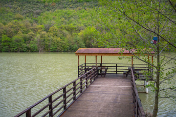 Obraz premium Wooden pier with gazebo over lake and forest backdrop. Serene lakeside nature spot. Rustic wooden canopy with red-roofed gazebo over calm lake waters and green hills. Relaxation concept
