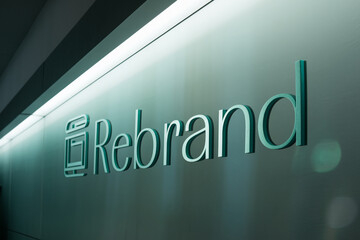 Teal "Rebrand" signage on a green wall, illuminated by overhead lighting, showcasing a modern and corporate design aesthetic, representing renewal and transformation in branding