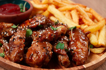barbecue chicken wings, sprinkled with sesame seeds, with French fries, and sauce, on a wooden plate, rustic style,