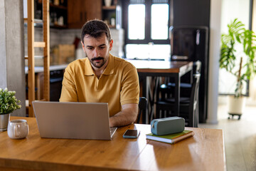 Young man concentrating on his work, typing on a laptop at the kitchen table in his modern apartment