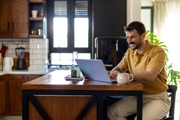 Smiling man working remotely using laptop in kitchen, enjoying flexible work schedule and work-life...