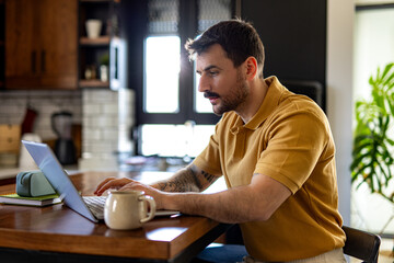 Young man concentrating while working on his laptop in a modern kitchen, enjoying a cup of coffee...