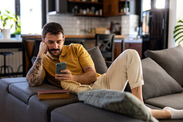 Obraz premium Relaxed young man lying on a comfortable sofa in a modern living room, using his mobile phone while enjoying his leisure time