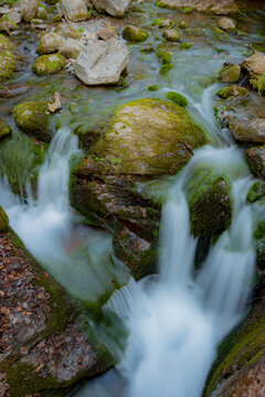 Imagen serena de un peque&ntilde;o riachuelo fluyendo entre piedras lisas, con el agua creando una apariencia sedosa y suave gracias al efecto de larga exposici&oacute;n. El entorno natural y tranquilo es ideal par