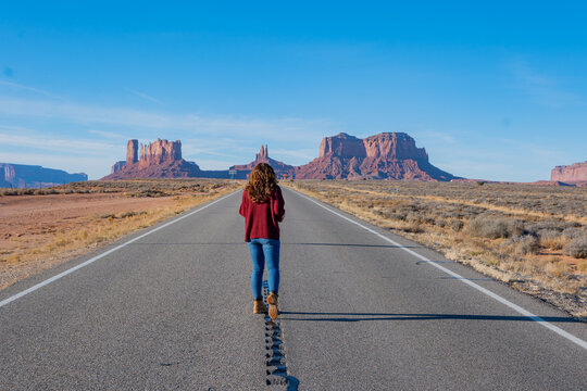 Una mujer vista de espaldas caminando por una larga carretera recta que se dirige hacia las formaciones rocosas ic&oacute;nicas de Monument Valley. El vasto paisaje des&eacute;rtico, junto al sentimiento de soledad