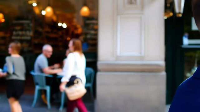 Young man in a suit listening to music with headphones while walking past a sunny outdoor caf&eacute;

