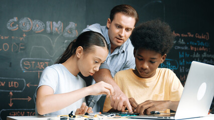 Caucasian teacher talking about electric tool while student fixing robotic model on table with electronic equipment placed. Diverse academic children learning about main board structure. Edification.