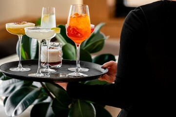 A server carries a tray with four colorful cocktails, including a spritz, margarita, and a creamy dessert drink. Lush green leaves in the background enhance the inviting atmosphere.
