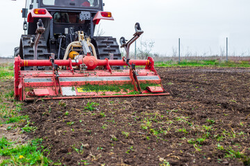 A tractor equipped with a red rotary cultivator actively prepares the soil in a rural field. The...