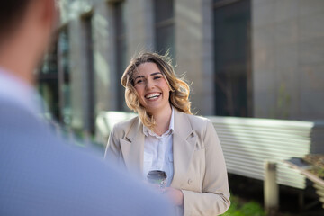 Smiling businesswoman holding a coffee cup and talking to a male colleague outdoors. Friendly professional interaction in a modern office district.