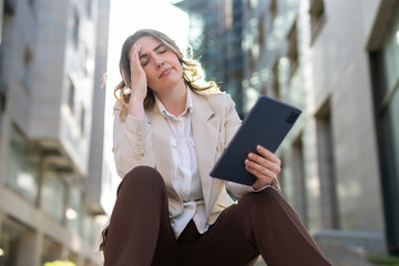 Frustrated businesswoman sitting outdoors in the city, holding a digital tablet and touching her head in stress. Young professional female facing technical problems or bad news at work.
