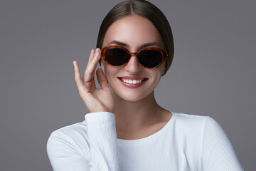 Eye wear fashion portrait of a young woman in oval tortoiseshell sunglasses and white ribbed top. She touches the frame with one hand, stylish and confident. Studio shot on gray background.