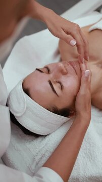 Young woman lying on bed at spa salon and enjoying pampering facial massage. Cosmetologist massaging face of female client for lymphatic drainage and lifting effect. Beauty, cosmetology, wellness