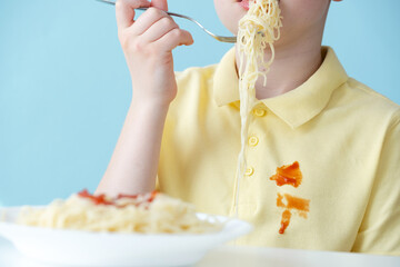 A child eats spaghetti with a fork while wearing a yellow shirt that has sauce stains. The setting...