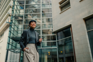 Smiling businesswoman holding tablet and looking up in modern office building exterior