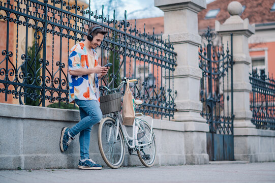 Stylish man listening to music on headphones and using smartphone while leaning on his bicycle