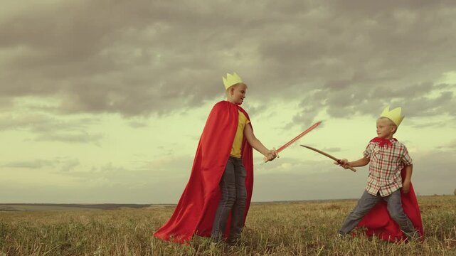 children superheroes play with swords field kings knights. dream childhood. little kid and his brother fearless defenders. teamwork child boys sons. dream adventure red capes at sunset. summer games