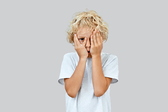Shy and curious, an 8 years old blonde boy with curly hair hides his face behind hands, peeking through fingers in a light t-shirt on grey background.