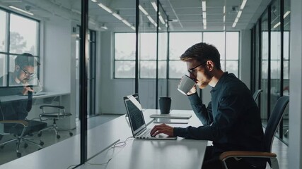 Professional young businessman multitasking, drinking coffee while working on laptop in bright, modern office interior with minimalist design and clean workspace