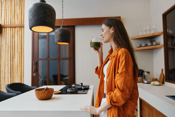 Young woman enjoying a healthy green smoothie in a bright, modern kitchen, dressed in an orange shirt and white dress, exuding a sense of serenity and wellness