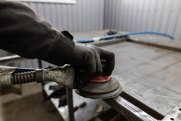 A worker's gloved hand uses a power sander to smooth the joints of a metal frame on a workbench in...