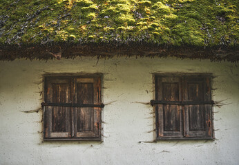 Part of an abandoned old traditional house with wooden windows and a slate roof covered moss and lichen. Two brown windows with closed shutters covered cobwebs on a white wall of a clay house