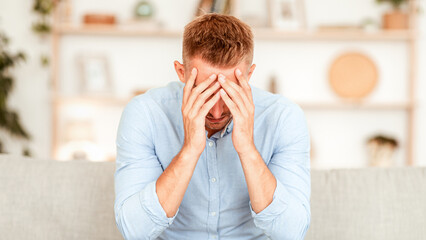 Depression Concept. Portrait of stressed man sitting alone on couch in living room, covering his face with hands, crying. Upset guy in despair thinking about problems or failure, copy space
