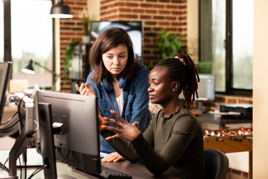 Two multiethnic female colleagues brainstorming and discussing creative ideas for a marketing agency. Caucasian manager focused as a black woman explains project updates from her desktop monitor.