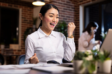 Closeup of asian female professional reacting with joy at her desk, raising her fists in excitement while looking at laptop screen. Her expression captures happiness, success, and sense of achievement