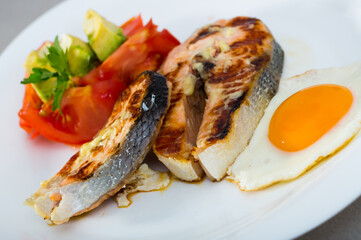 Grilled salmon served with fried egg, fresh tomatoes, avocado and parsley on white dish..