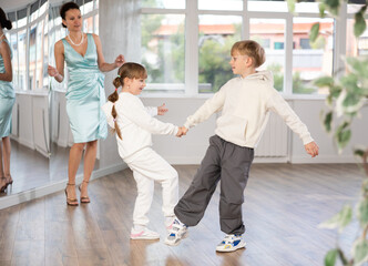 Fototapeta premium Boy and girl kids repeat movement while taking boogie woogie dancing lessons. Female teacher conducts class for students during dance workout and watches performing movements