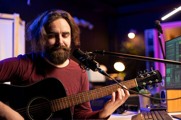 Obraz premium Skilled composer playing acoustic guitar and singing on microphone in home studio, mixing and mastering songs with electronic tools. Music producer recording tunes on daw software.