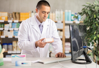 Fototapeta premium In utility room of pharmacy, male pharmacist works at computer and pill box product, reads drug database. Tracking movement of drugs, ordering from supplier.
