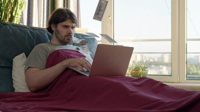 Freelancer man making money online using laptop, lying on bed, earn money from the comfort of bed. Cash flow. Money rain flow through the laptop. Dollar bills flying in investor face. Good investment