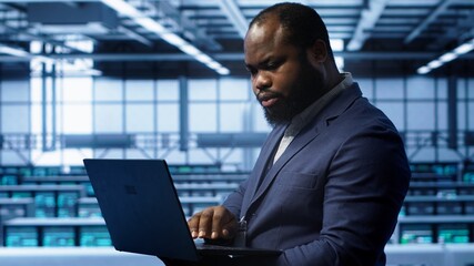 Fototapeta premium Software developer in server hub checking recovery plan on laptop, monitoring hardware energy consumption. African american worker uses notebook in data center, ensuring rigs are functioning, camera A