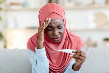 Sick black muslim woman in hijab looking at thermometer at home, checking temperature, suffering...
