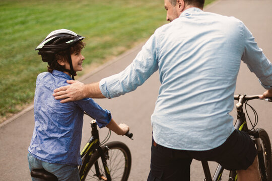Healthy Family Relationship. Rear back view portrait of smiling father and son riding bicycles down the street in public park, man tapping and patting boy on the shoulder, teaching and showing support