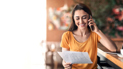 Happy arab lady having conversation on mobile phone at cafe, holding documents, working online, copy space. Smiling woman talking on smartphone with clients while drinking coffee, looking at papers