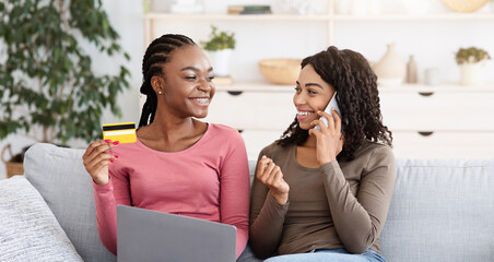 Black cheerful girlfriends ordering food online, making confirmation on smartphone, holding laptop...