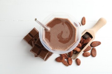 Tasty chocolate milk, bars and cocoa beans on white wooden table, flat lay
