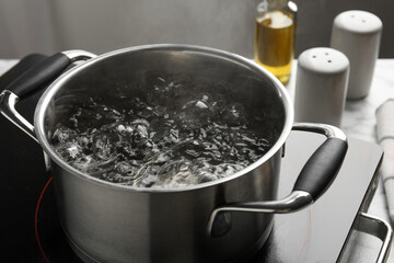 Cooking pot with boiling water and stove on white table against grey background, closeup