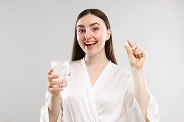 Happy woman with glass of water and collagen pill on grey background