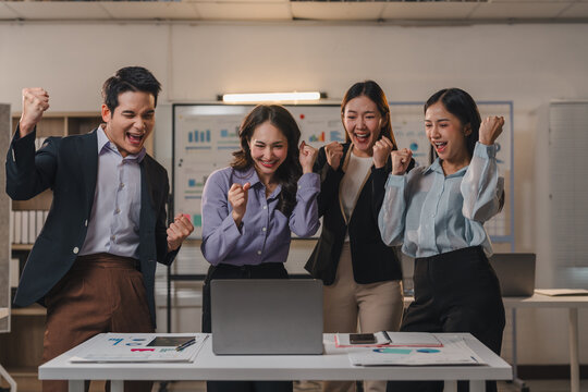 Four enthusiastic asian businesspeople are cheering and raising their fists in front of a laptop in a modern office, expressing joy and teamwork after achieving a successful business outcome
