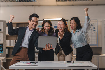 Group of happy and enthusiastic asian businesspeople celebrating a successful project, raising their arms in joy and looking at a laptop in a modern office environment