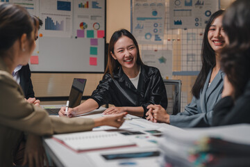 Group of young asian businesswomen happily discussing and working together during a meeting with financial charts and graphs displayed on a whiteboard in a modern office